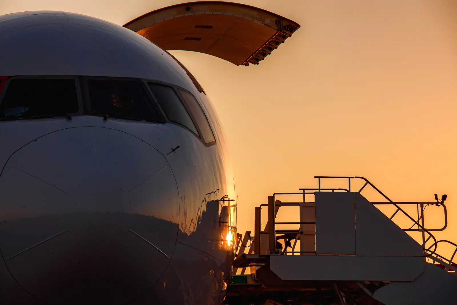 The nose of a cargo aircraft with its door raised and a loading platform, at sunset
