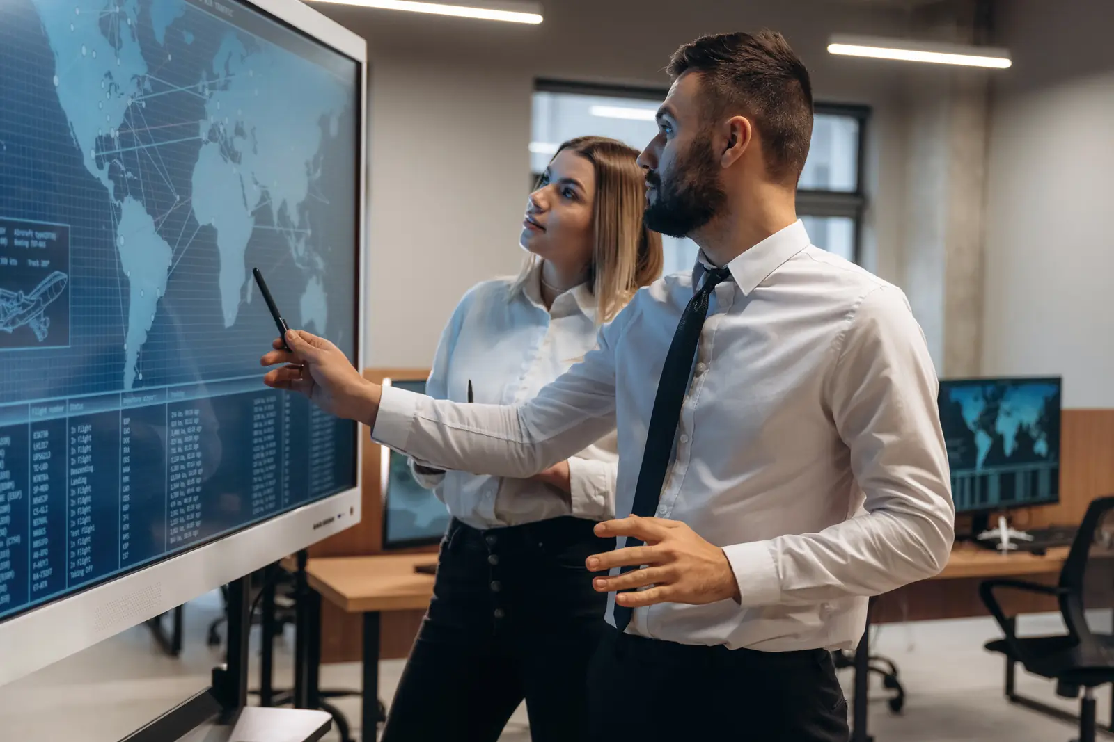 Two transport coordinators studying a world map with routes on a large screen in an office