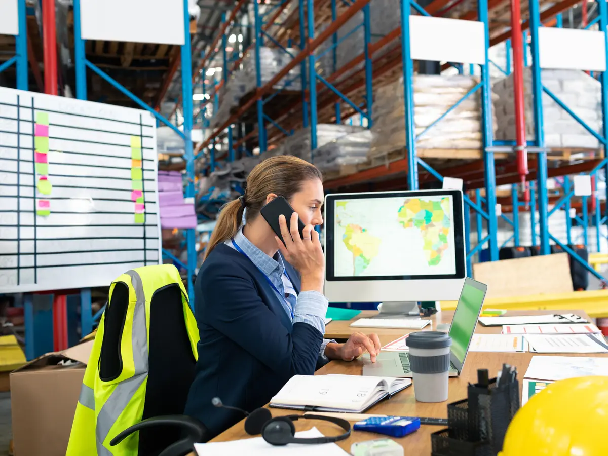 A dispatcher on the phone at a desk in a warehouse, with a world map on the monitor