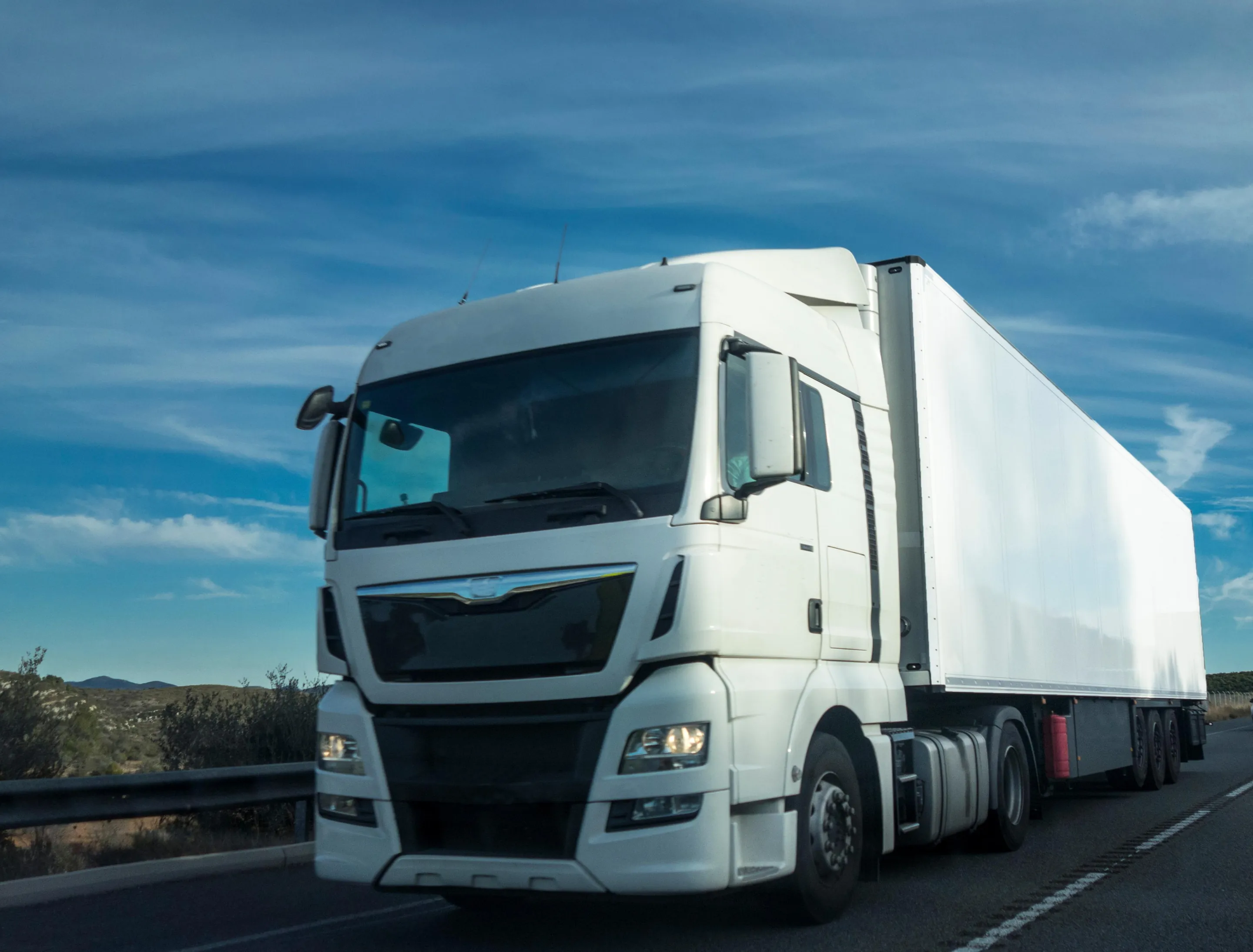A European tractor unit with a white refrigerated semi-trailer, driving on a motorway under a blue sky