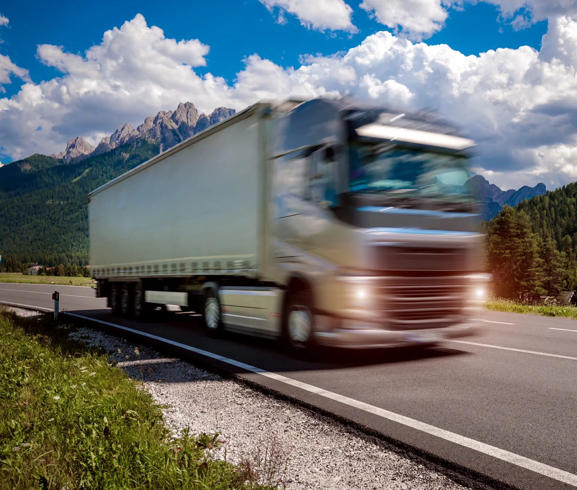 A modern European truck driving along a mountain road towards the viewer, with the Alps in the background