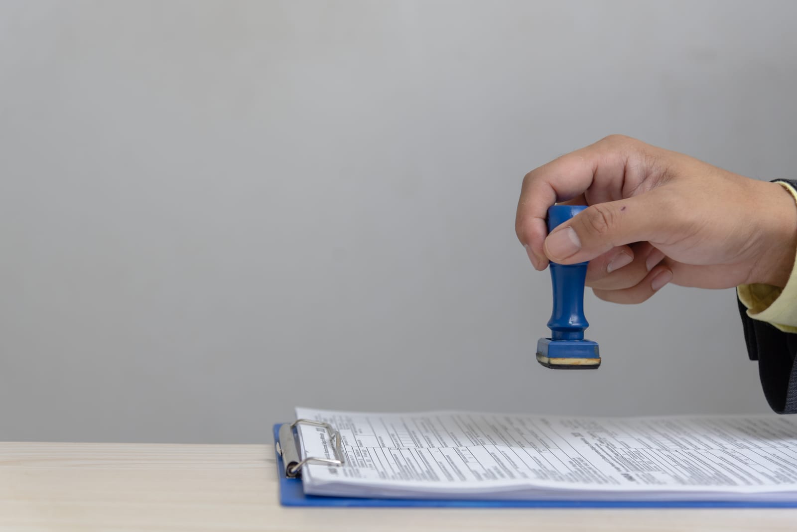 Mână care aplică o ștampilă pe documente prinse într-o planșetă albastră, pe un birou