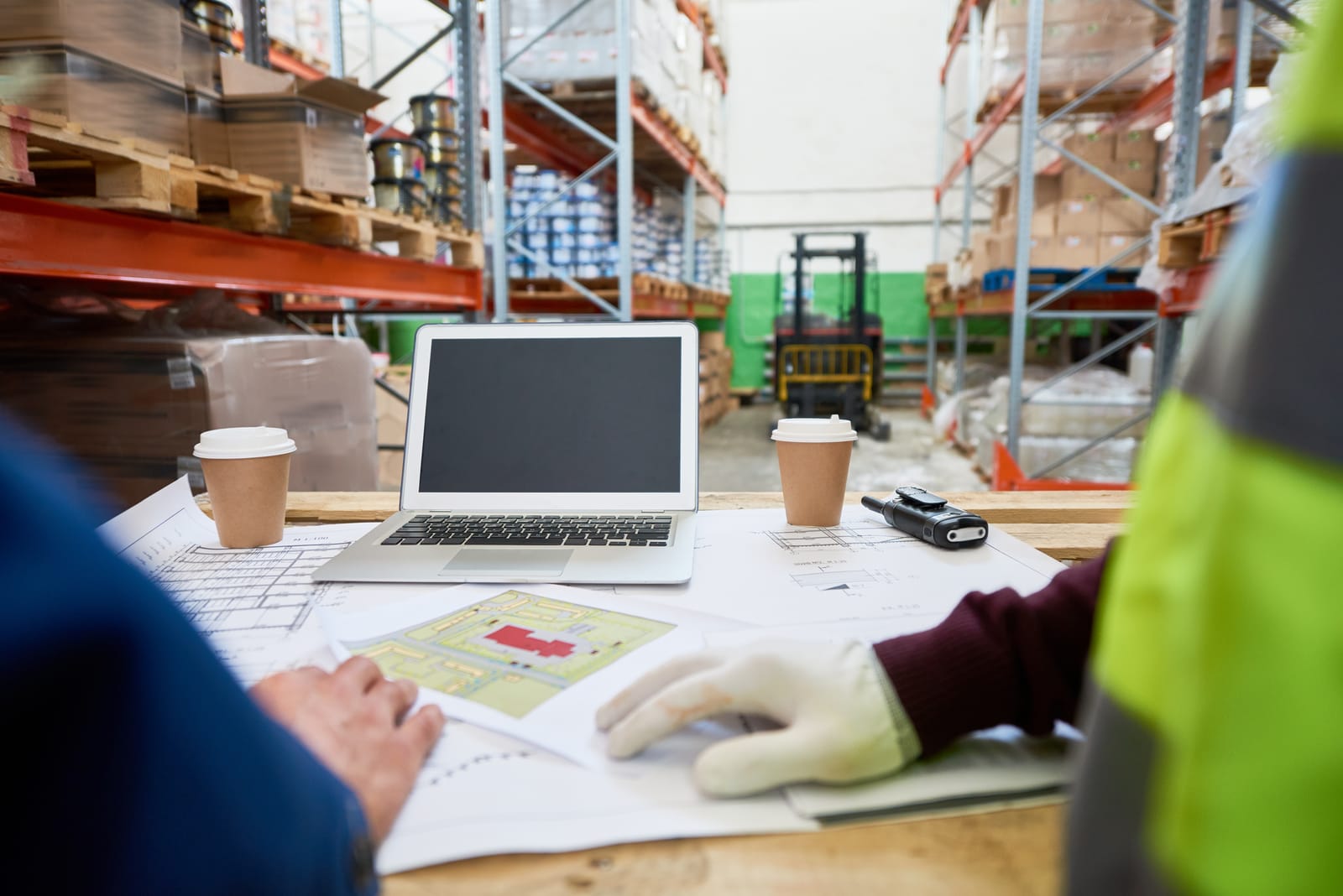 Laptop, plans and coffee cups on a table, in front of warehouse racking