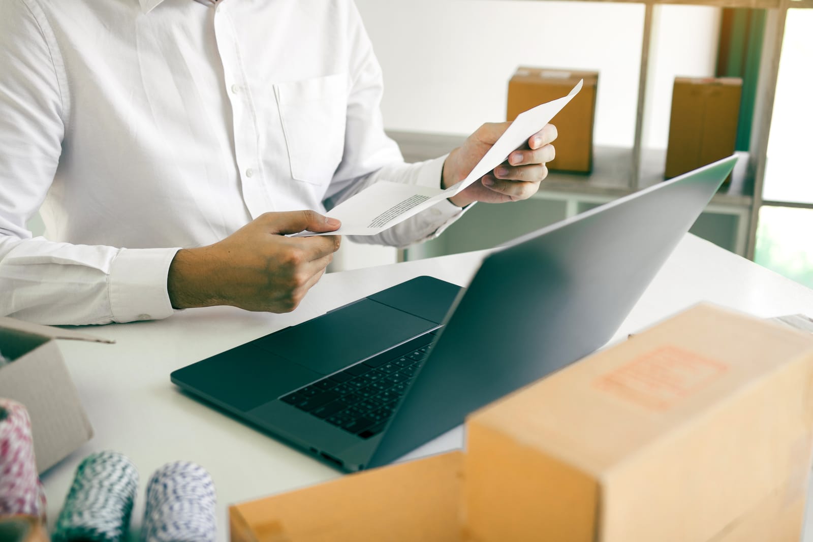 Man in a white shirt checking documents next to a laptop, in front of some cardboard boxes