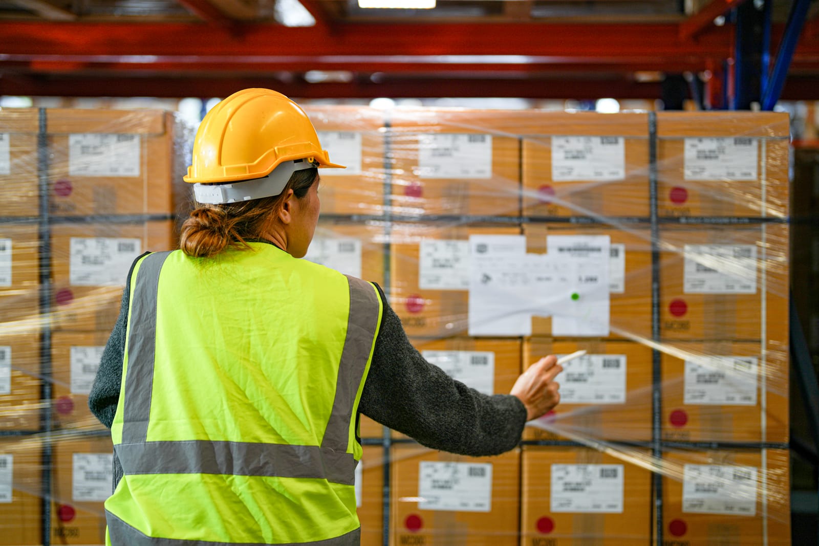 Female worker in a high-visibility vest and hard hat checking the labels on palletised, film-wrapped packages