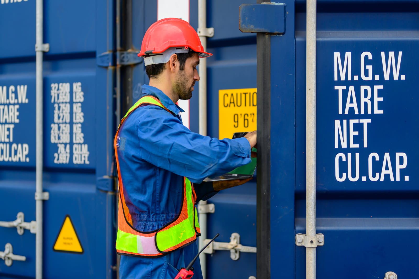 Worker in a high-visibility vest inspecting a freight container, holding a laptop