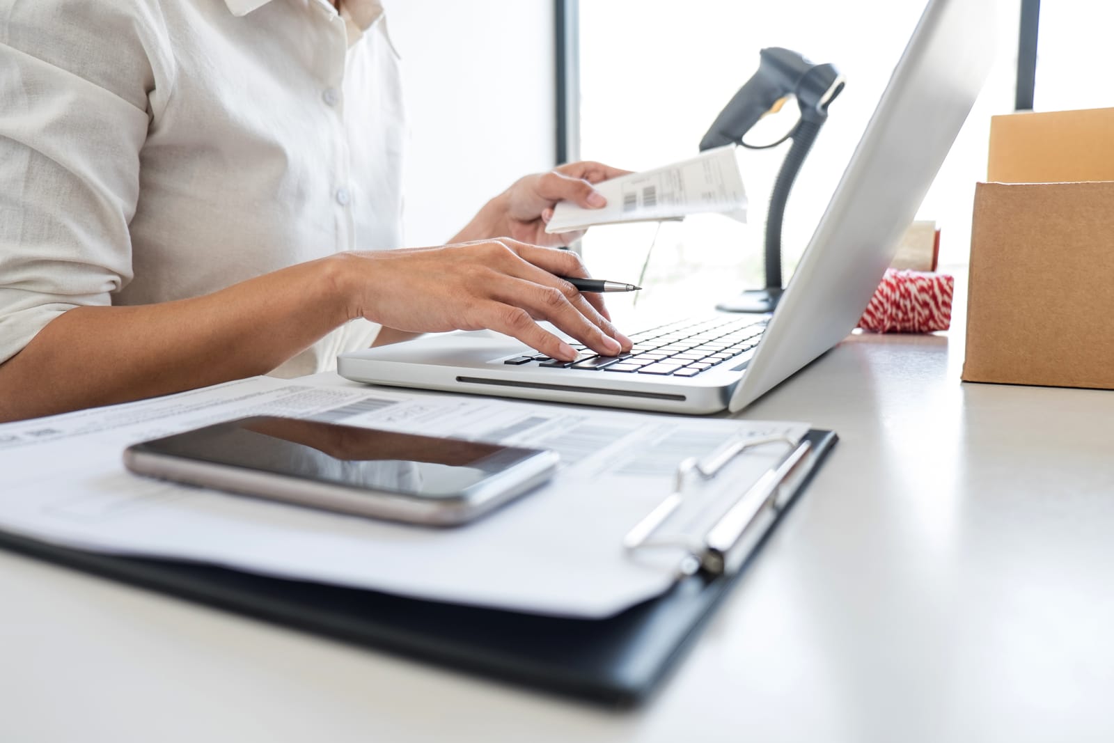 Person working on a laptop, with a barcode scanner, a phone and a folder of documents on the desk